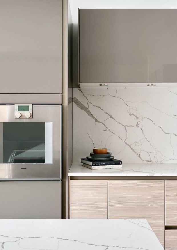 Close-up of a refined contemporary kitchen featuring integrated stainless-steel appliances, muted taupe upper cabinetry, reeded light-wood lower cabinetry, and a dramatic white marble-look stone splashback and island benchtop with grey veining. A small coffee cup and books add minimal styling.