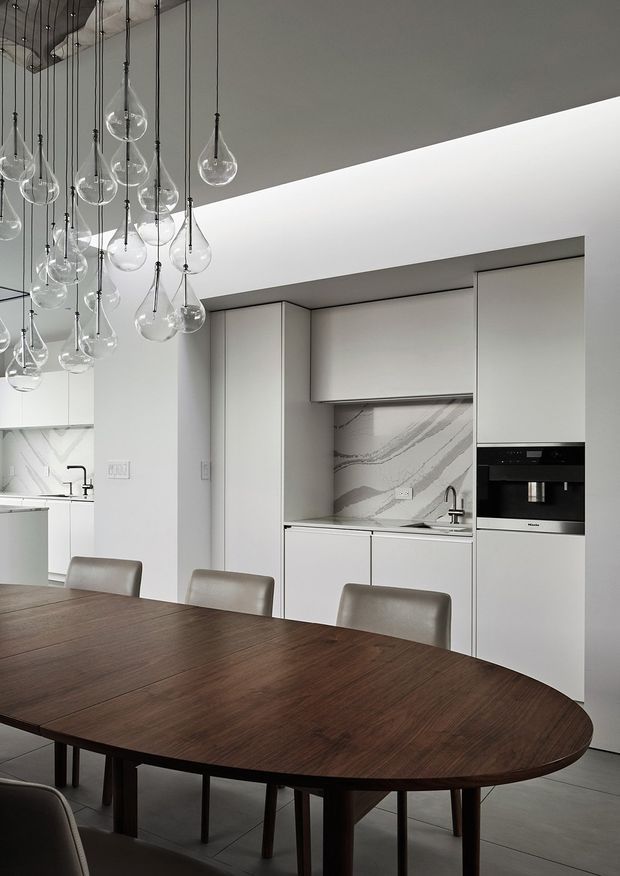 A refined contemporary kitchen and dining area with a large dark timber oval dining table in the foreground, white handleless cabinetry, integrated black appliances, and a dramatic marble-look splashback. Numerous clear glass globe pendant lights hang at varied heights from the ceiling, creating a sculptural focal point. The palette is predominantly white, grey, and dark timber with a clean, restrained finish.
