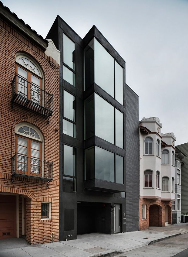Street-level exterior view of a narrow multi-storey urban infill building with a dark contemporary facade, large vertically arranged windows, and projecting angular volumes. It is tightly flanked by ornate brick and rendered heritage-style neighbouring buildings, creating a strong contrast between modern and traditional architecture.
