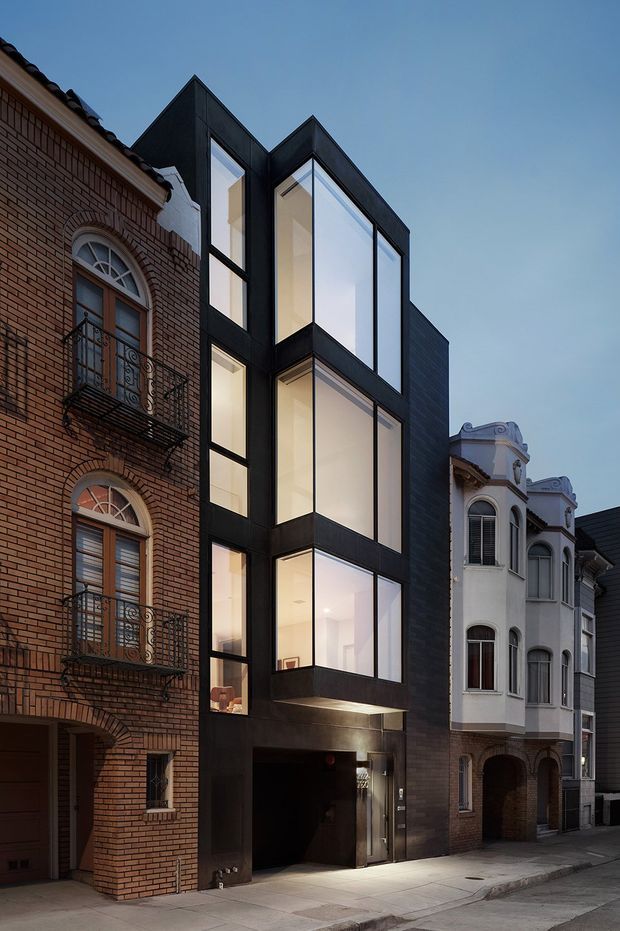 Street-level view of a narrow contemporary multi-storey infill building between ornate historic brick and rendered townhouses. The new facade is clad in dark panels and composed of projecting angular volumes with extensive floor-to-ceiling glazing, illuminated interiors, and a recessed ground-floor entry or garage opening. The surrounding streetscape includes heritage-style arched windows, balconies, decorative cornices, and a paved sidewalk at dusk.
