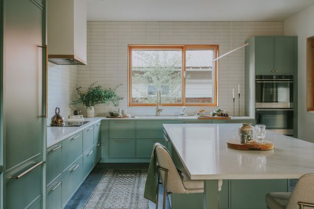 A bright, spacious contemporary kitchen with muted sage-green cabinetry, white tiled walls, warm timber window framing, and a large pale stone island with integrated seating. The L-shaped kitchen includes a stainless-steel sink beneath a timber-framed window, full-height integrated appliances, white benchtops, and a patterned runner rug. The styling is calm and refined with natural greenery and warm neutral accents.