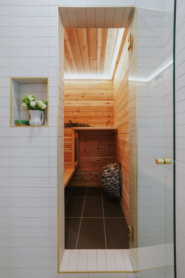 A compact sauna viewed through an open glass door, with warm timber wall and ceiling panelling, built-in wooden bench seating, a dark tiled floor, and a metal sauna heater basket filled with stones. The surrounding room is clad in white rectangular tiles with a recessed display niche and brass-toned trim and hardware.