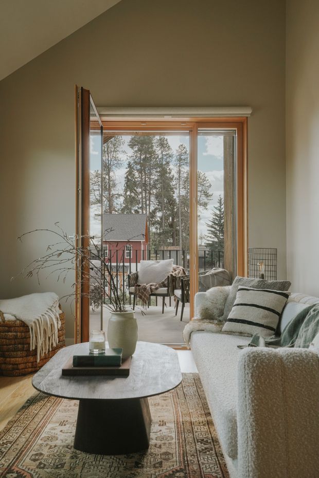A warm, cozy living room with a large timber-framed glazed door opening to a small balcony or terrace. The room features a textured light sofa, woven throw, patterned area rug, rounded coffee table, woven accent seating, and a neutral olive-beige wall palette. Tall trees and a red house are visible outside through the glazing.