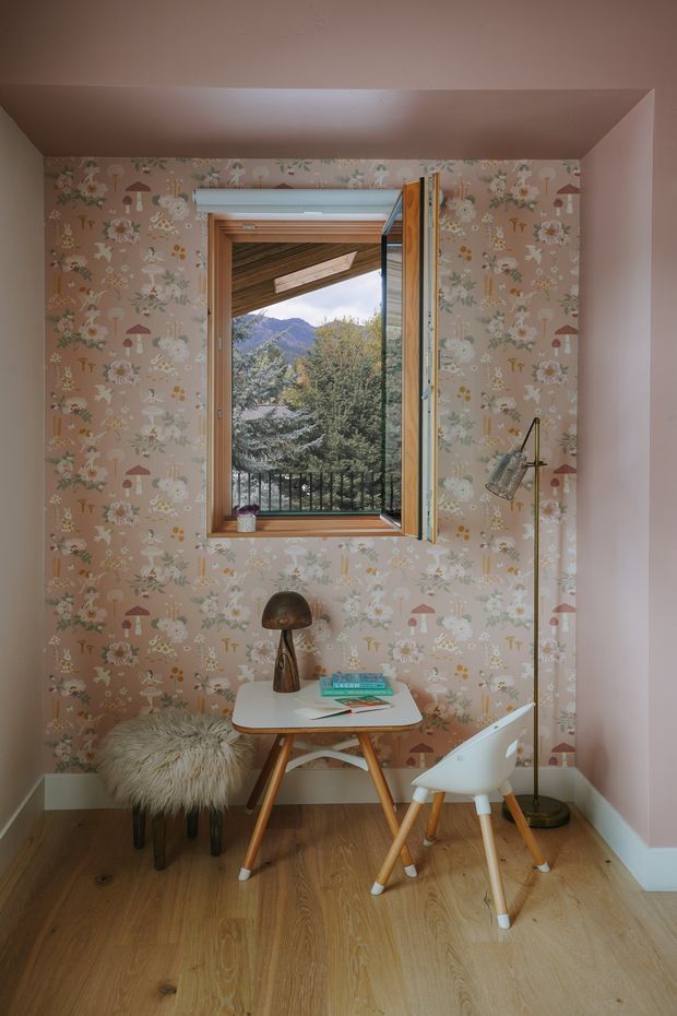 A small, warmly styled home office or study nook with a compact timber-legged desk, white chair, furry stool, desk lamp and mushroom-shaped table lamp. The room features playful pink floral-and-mushroom wallpaper, timber flooring, pale pink painted walls and a timber-framed opening window. Through the window, a covered exterior structure, trees and mountains are visible.