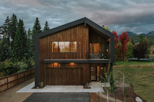 Exterior view of a compact two-storey timber-clad house in a mountainous, wooded setting. The home has a steep gable roof with deep dark trim, a projecting upper-level balcony, large glazed openings, and an integrated ground-floor garage. Dark weathered wood contrasts with black framing and concrete paving.
