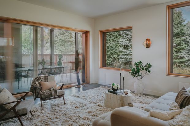 Bright, softly furnished living room with large timber-framed windows and sliding glass doors overlooking mature trees. The room includes a cream sectional sofa, patterned lounge chairs, a shag rug, a round stone or concrete coffee table, and a sculptural wall sconce. A dining or outdoor seating area is visible beyond the glazing.