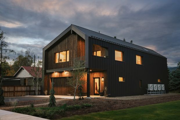 A modern two-storey detached house photographed at dusk. The dark, gable-roofed building has a vertically ribbed dark metal-clad facade, warm timber or wood-toned recessed sections, large illuminated windows, and a sheltered entry. It sits within a landscaped suburban setting, with a smaller older structure visible behind it.