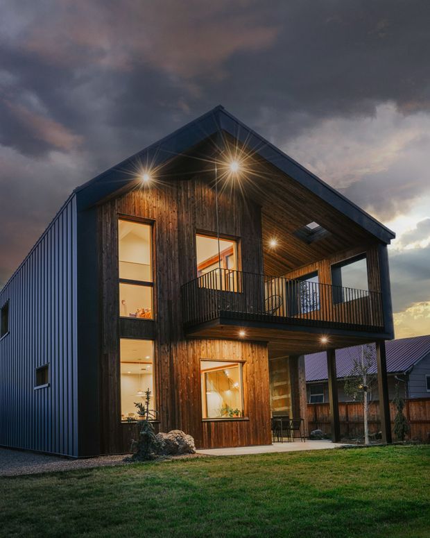 Exterior photograph of a contemporary two-storey gabled house at dusk. The facade combines dark vertical timber cladding with dark corrugated metal side walls and roof-like gable framing. Large glazed openings, an upper covered balcony, black metal balustrades, exposed timber soffits, and warm exterior lighting define the elevation. The home sits in a suburban residential setting with lawn and neighbouring houses visible.