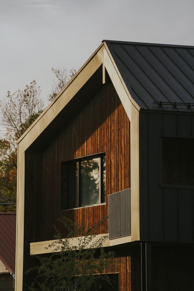 Close-up exterior view of a compact modern dwelling with a steep gabled roof. The facade combines dark vertical timber cladding with black metal roofing and dark metal-framed glazing, accented by pale trim around the gable. A small upper balcony with slender metal balustrading is visible, with trees and neighbouring rooflines in the background.