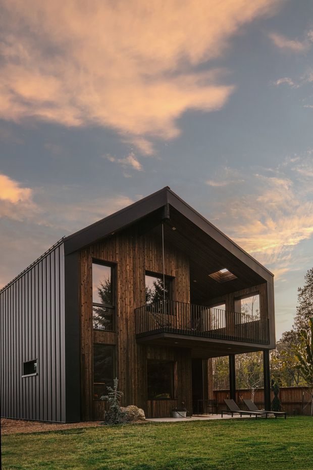 Exterior view of a contemporary two-storey gable-roof house at dusk. The building combines dark vertical metal cladding with a weathered timber-clad gable facade, expansive dark-framed glazing, a deep covered upper balcony, and a sheltered ground-level outdoor living area. The house sits within a landscaped lawn and wooded setting beneath a dramatic sunset sky.