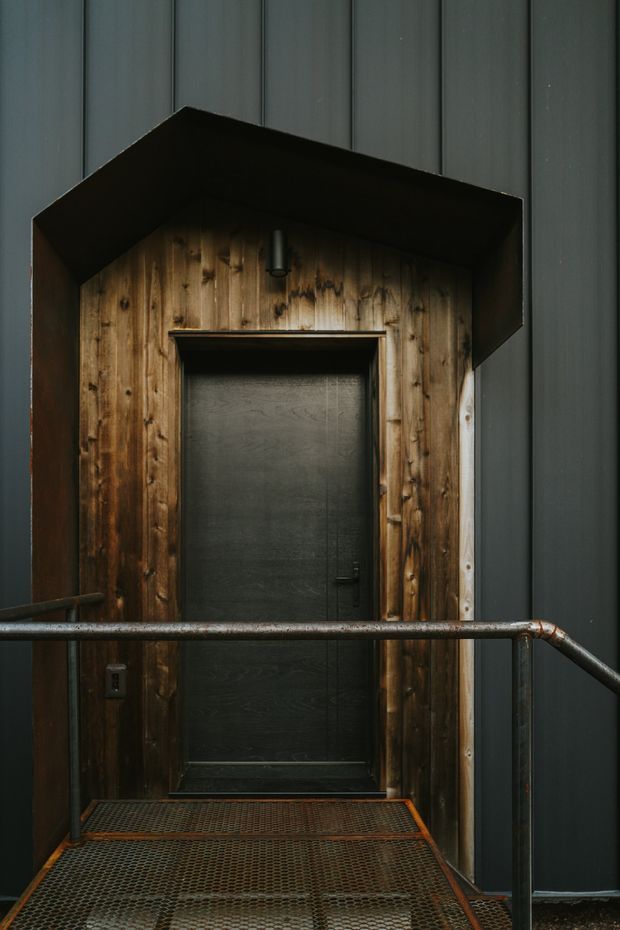 A moody industrial-style exterior entrance with a dark metal-clad facade, timber-lined doorway, black door, projecting metal canopy, and weathered steel mesh landing with tubular handrails.