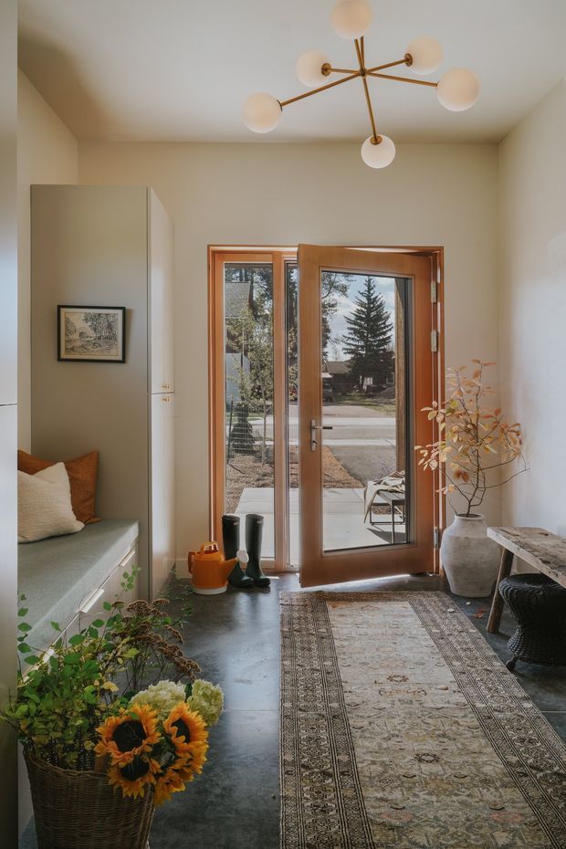 A warm, softly styled residential entryway viewed from inside. The space features a wood-framed glazed front door with sidelights, pale neutral walls, a dark polished concrete floor, a built-in bench with cushions on the left, a patterned runner rug, a sculptural multi-arm pendant light, and a console table with a large ceramic planter. Natural light enters from the street-facing glazing, creating an inviting transition between indoors and outdoors.