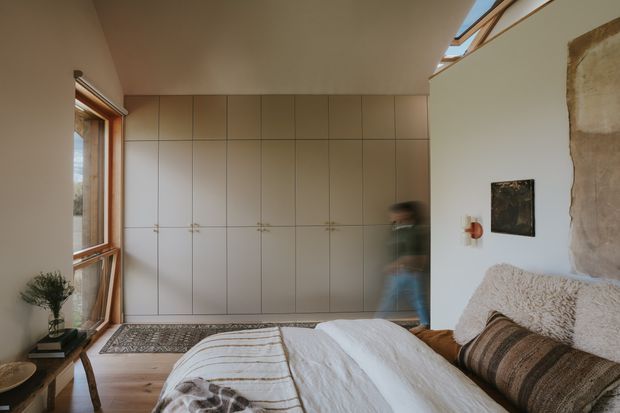 A warm, minimalist bedroom with a low bed in the foreground, a full wall of pale built-in wardrobes, timber-framed windows, a skylight, and a partially exposed textured wall. Natural timber flooring, layered neutral bedding, and soft wool textiles create a calm, earthy atmosphere.