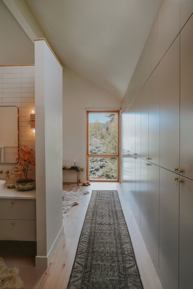 A narrow, warm-toned interior passage beside a bathroom or vanity area, with full-height pale built-in cabinetry lining the right wall, a tall timber-framed window at the end, and light timber flooring partly covered by a long patterned runner. On the left, a tiled wet-area wall, vanity, mirror, wall light and indoor plants are visible. The space has a sloping ceiling and soft natural light, creating a calm, refined atmosphere.