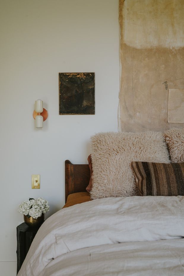A close-up of a warmly styled bedroom corner featuring a wooden bed with layered cream bedding, a shaggy cream throw, and textured striped cushions. The pale painted wall includes a small artwork and sculptural wall sconce, while a dark bedside table holds a vase of flowers. A large raw, aged plaster or canvas-like wall treatment appears behind the bed, creating a tactile, earthy contrast.