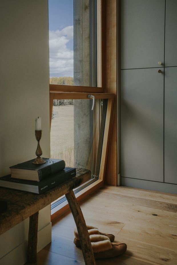 A warmly lit interior corner featuring a tall timber-framed window, pale timber flooring, and a large flush grey built-in storage cabinet with small round pulls. A rustic timber console table holds stacked books and a candle, while soft slippers rest on the floor. The window reveals a rural landscape and contributes natural light to the calm, understated space.
