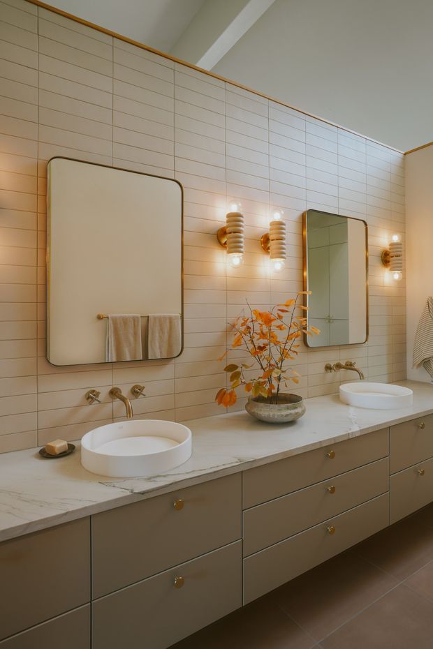 A warm, refined bathroom with a long double vanity, two white vessel basins, pale stone countertop, taupe built-in drawers with brass knobs, and brushed brass wall-mounted tapware. The walls are clad in horizontal cream tiles, complemented by rounded rectangular mirrors and sculptural ribbed wall sconces. A small decorative branch arrangement adds an organic accent.