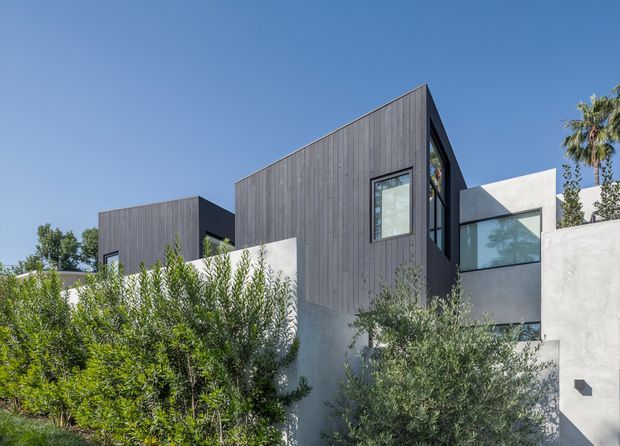 Exterior view of a contemporary multi-level residence with a strongly geometric composition. Dark vertical timber cladding forms box-like upper volumes, contrasted with smooth white rendered walls and large modern glazed openings. Dense Mediterranean-style planting partially screens the lower facade, with a clear blue sky and palm visible in the background.