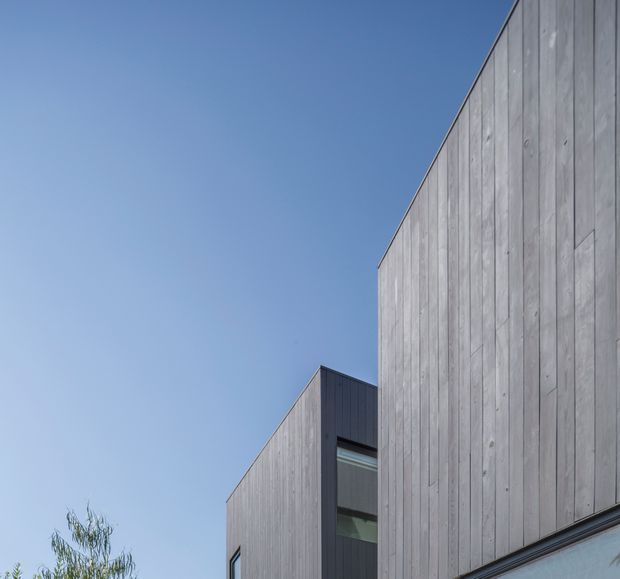 Close-up exterior architectural view of a modern building composed of tall vertical grey timber-clad volumes with crisp geometric rooflines and dark-framed glazing. A clear blue sky and a small amount of foliage appear in the background.