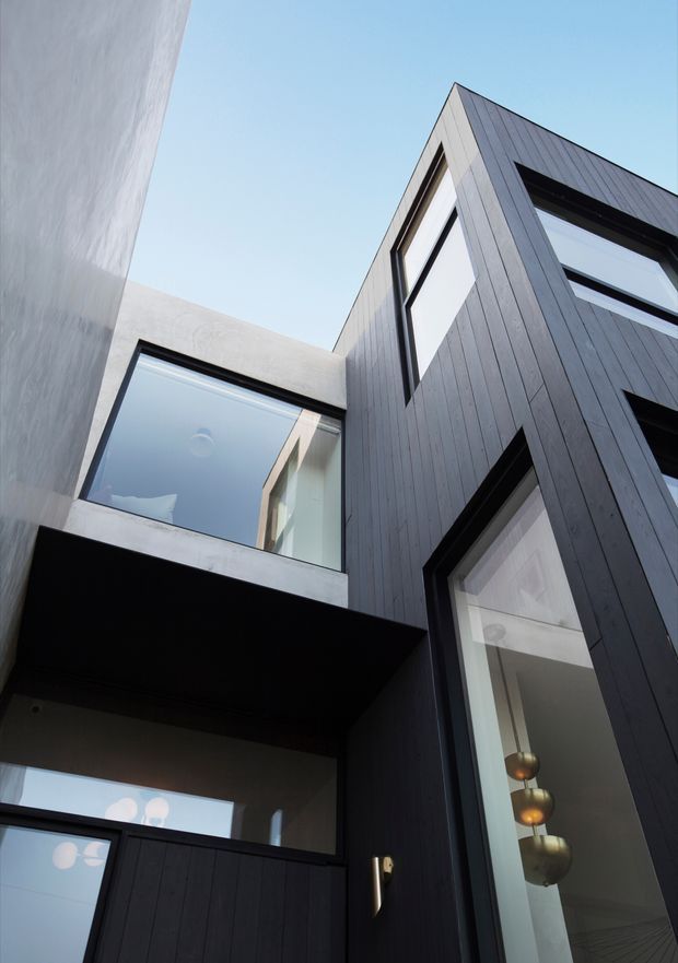 Upward-looking view of a contemporary multi-storey residential facade enclosing a narrow courtyard or entry void. The building combines dark vertical timber cladding, pale rendered or concrete walls, strong geometric projections, and large black-framed windows. Interior pendant lights are visible through the tall glazing, while the composition emphasizes the angular roofline, vertical proportions, and courtyard-like space.