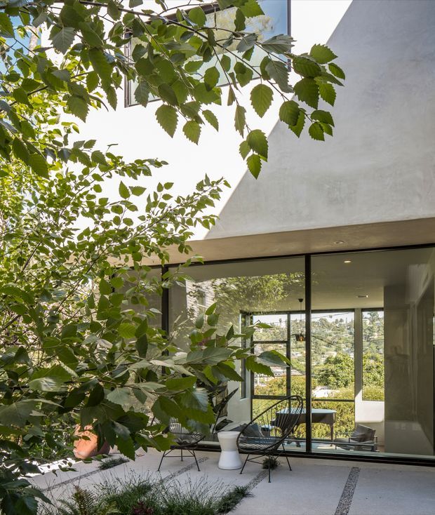 A contemporary courtyard or patio framed by leafy planting, with a concrete exterior wall and broad floor-to-ceiling glazed doors opening into a bright interior. Minimal black-framed glazing, a pale paved floor with narrow dark gravel or stone bands, and lightweight outdoor lounge chairs create a serene indoor-outdoor setting. A leafy suburban landscape is visible through the glazing.
