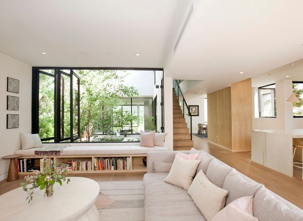 A bright, open-plan contemporary living area with a large curved sectional sofa facing a low built-in timber bookshelf and window seat. Full-height black-framed glazing opens toward a leafy courtyard, while a timber staircase, pale timber wall joinery, and a light kitchen island are visible in the background. The space has a clean, refined, natural palette with abundant daylight.