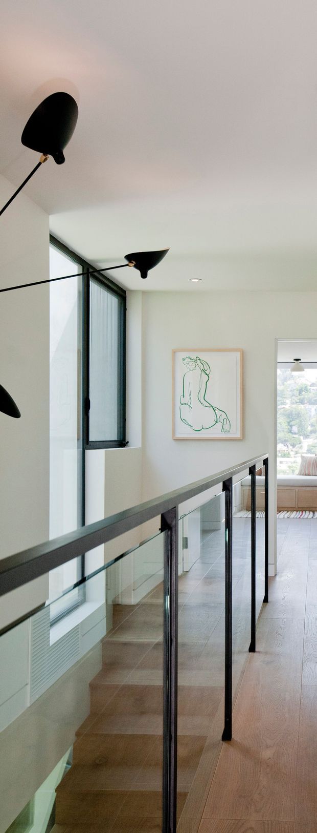 A bright, minimalist residential landing overlooking a staircase. The space features pale walls and ceiling, light timber flooring and stair treads, a slim black metal and clear-glass balustrade, tall black-framed glazing, and sculptural black wall-mounted light fixtures. A framed line drawing hangs on the wall, with a glimpse into an adjoining bright room beyond.