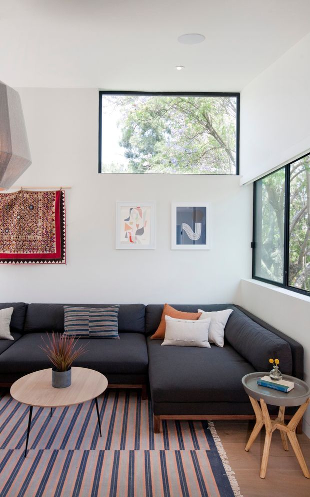 Bright contemporary living room with a dark charcoal L-shaped sectional sofa, striped area rug, round timber coffee table, and small side table. White walls and ceiling are paired with large black-framed windows overlooking trees, while a colourful woven wall hanging and framed abstract artwork add character. The room has abundant natural light and a clean, relaxed feel.