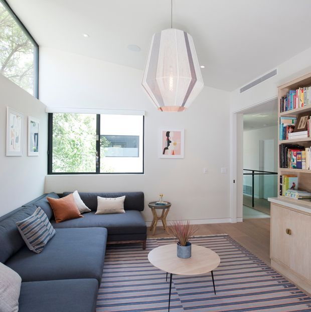 Bright contemporary living room with a large dark blue sectional sofa, striped area rug, round light-wood coffee table, sculptural pendant light, framed artwork, and a built-in timber bookshelf and cabinet. Large windows bring in abundant natural light, while a glazed hallway or stair area is visible through the opening at right.