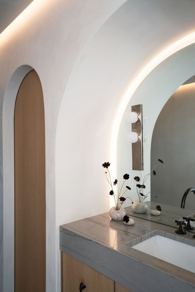 A refined bathroom vanity in a softly curved, arched interior. The space features a pale marble or stone benchtop with integrated rectangular basin, timber-look vanity cabinetry, a large arched mirror with concealed LED illumination, and sculptural globe wall lights. A tall arched timber door and curved plaster walls reinforce the organic architectural detailing. The palette is warm white, light timber, and pale grey stone, with dark floral stems as subtle decor.