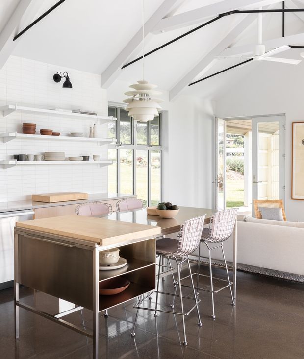 Bright open-plan kitchen and dining area with a large timber-topped island, integrated open shelving, dining table, bar stools and a partially visible living area. The room has white tiled walls, polished dark flooring, exposed white rafters, extensive glazing, and distinctive layered pendant lighting with a large ceiling fan. The palette is predominantly white, light timber and dark grey, creating an airy contemporary interior.