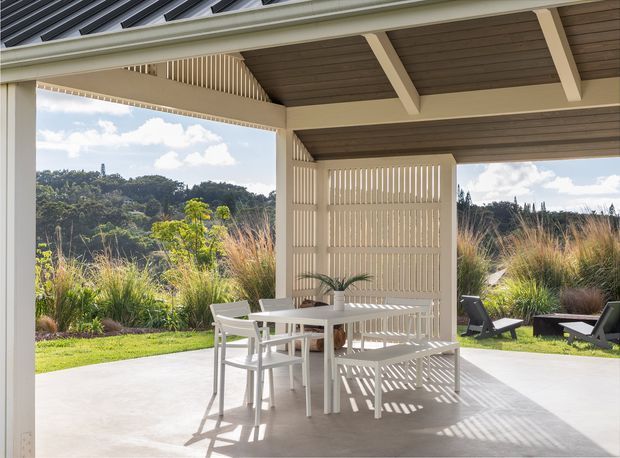 A bright, covered outdoor dining and lounge area with a white table, chairs, and built-in bench beneath a timber-lined pitched roof. Vertical slatted screens provide partial enclosure and cast patterned shadows across the concrete floor. The space opens onto landscaped planting and a green hillside, creating a contemporary indoor-outdoor setting.