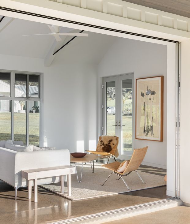 Bright, airy living room viewed through a large open glazed sliding door. The room has white walls, exposed white structural beams, a ceiling fan, polished warm-toned concrete flooring, a neutral rug, a white sofa, light timber and leather lounge chairs, a small coffee table, and framed botanical artwork. Large windows and glazed doors provide views to a lawn and neighbouring property.