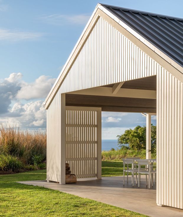 Exterior view of a minimalist coastal pavilion or small gabled house clad in pale vertical timber battens. The open-sided ground level forms a covered outdoor dining area, with exposed timber structure, a dark pitched metal roof, concrete paving, and views toward the ocean and landscaped grasses. The image has bright natural light and a clean, contemporary coastal character.