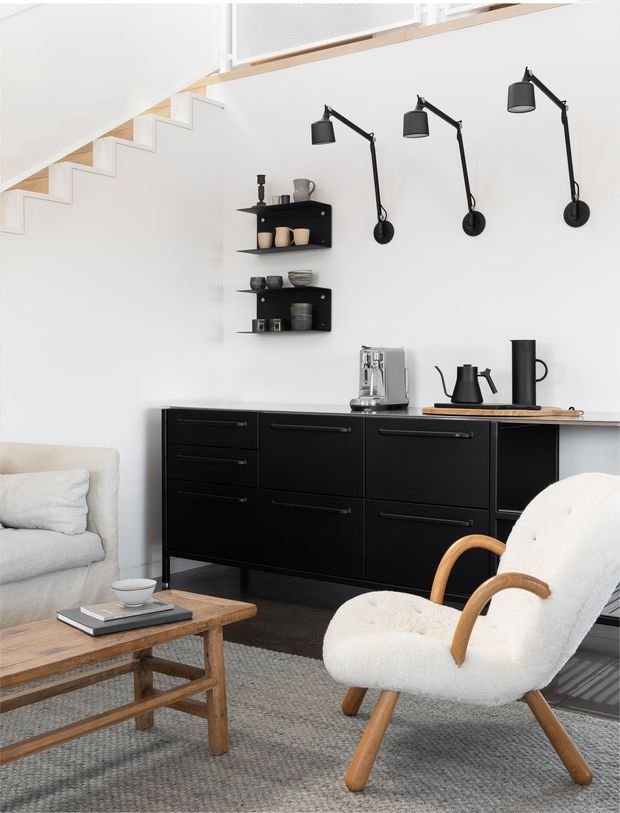 Bright, minimalist living area with a black built-in sideboard and open shelving, coffee-making station, sculptural wall-mounted lamps, pale upholstered seating, and a timber coffee table. A modern staircase with light timber detailing is visible above, and the room has a restrained black, white, grey, and natural-wood palette.