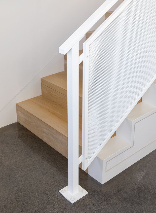 Close-up interior view of a modern staircase with warm light timber treads, white painted stringers and handrails, and a perforated white metal balustrade panel. The stair base incorporates white built-in cabinetry or drawers, while the surrounding floor is dark grey carpet. The design is clean, minimalist, and highly detailed.