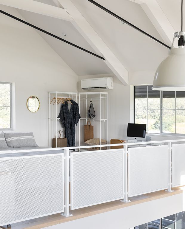 Bright, compact mezzanine-style home office or workspace overlooking a lower level, with white perforated metal balustrade panels, a white desk and computer, open white clothing/storage racks, a grey sofa, large windows, and exposed white structural ceiling beams. A large white pendant light and black linear ceiling details add contrast. The room has a clean, airy contemporary feel.