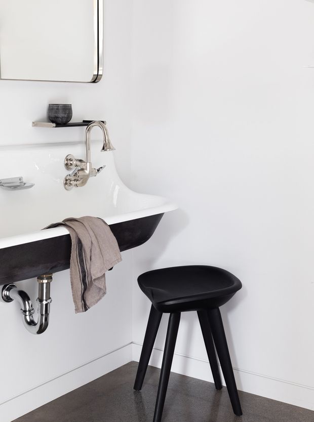 A minimalist bathroom vignette featuring a wall-mounted white porcelain basin with a contrasting black underside, exposed chrome plumbing, and a polished chrome wall-mounted faucet. A black stool sits beside the basin, with a neutral taupe towel draped over the edge. The space has plain white walls, simple white skirting, and a dark grey floor covering. A rounded rectangular mirror and small shelf with a dark vessel are partially visible above the basin.