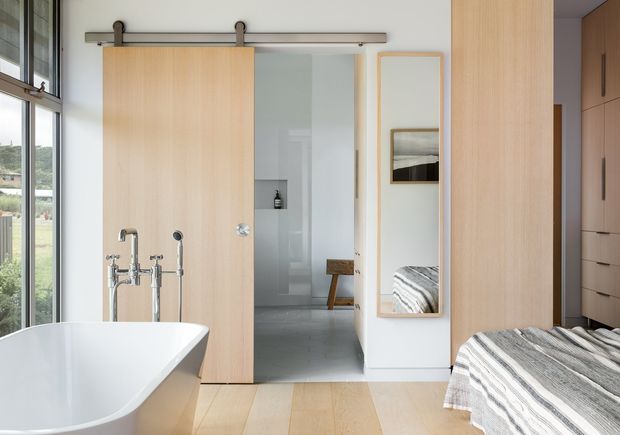 Bright contemporary bedroom with an integrated freestanding bathtub in the foreground, separated from the sleeping area by a large pale timber sliding door. The room features pale timber flooring and cabinetry, white walls, a full-height window, a wall-mounted mirror, and a partially visible bed with striped bedding. A grey-tiled bathroom or wet area is visible through the opening beyond the sliding door.