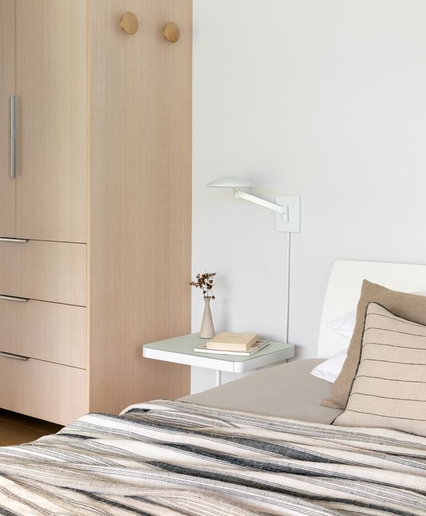 Bright, refined bedroom interior featuring a light timber built-in wardrobe with integrated drawers and simple round knobs, a floating bedside table, a minimalist wall-mounted reading lamp, and a neatly made bed with neutral linen bedding and a striped throw. The palette is soft and natural, with clean contemporary detailing and abundant white wall space.