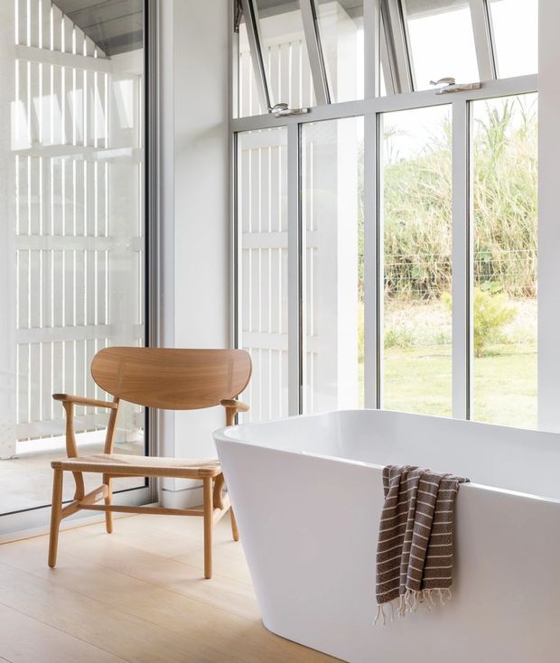 Bright, airy contemporary bathroom featuring a freestanding white bathtub positioned beside extensive floor-to-ceiling glazing. A sculptural timber lounge chair and white vertical privacy screens frame the space, with pale timber flooring and a neutral palette. The glazing looks onto grasses and landscaping outside.