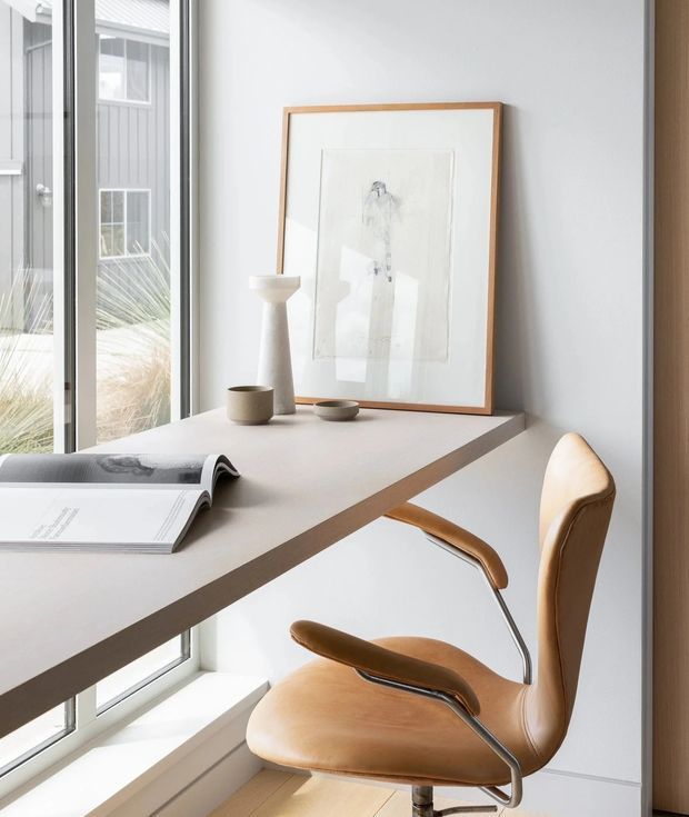 Bright minimalist home office nook beside full-height glazing, featuring a slim light-toned desk, sculptural framed artwork, simple ceramic accessories, and a tan leather swivel chair with metal arms. The space has pale walls, a light timber floor, and a clean, restrained contemporary aesthetic.