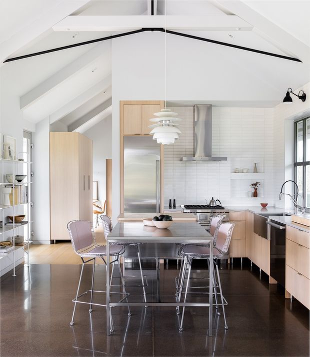 Bright open-plan kitchen and dining area with a vaulted white ceiling and exposed beams. The kitchen features light timber cabinetry, a white tiled splashback, stainless-steel appliances and rangehood, a central island with a pale benchtop, and a large stainless-steel dining table surrounded by translucent pinkish chairs and stools. Black polished flooring contrasts with the predominantly white and light timber palette. A tall glazed window and open shelving add to the airy contemporary interior.