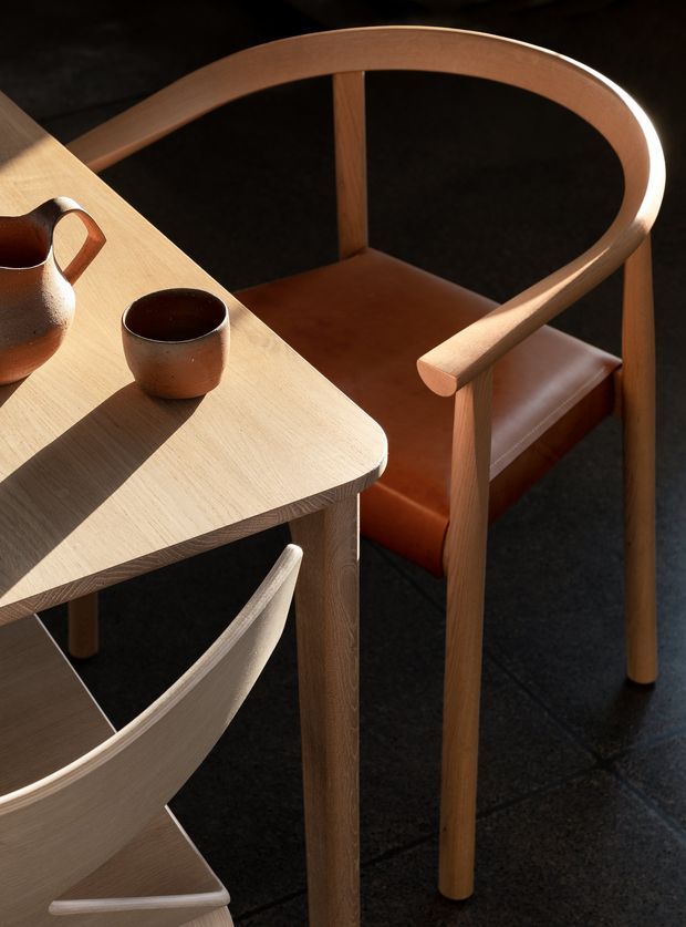 Close-up interior view of a refined dining setting with a light timber table and sculptural timber dining chairs. One chair has a rounded bentwood frame and warm brown leather seat and back, while ceramic vessels sit on the tabletop. Strong sunlight creates defined shadows across the table and dark tiled floor.