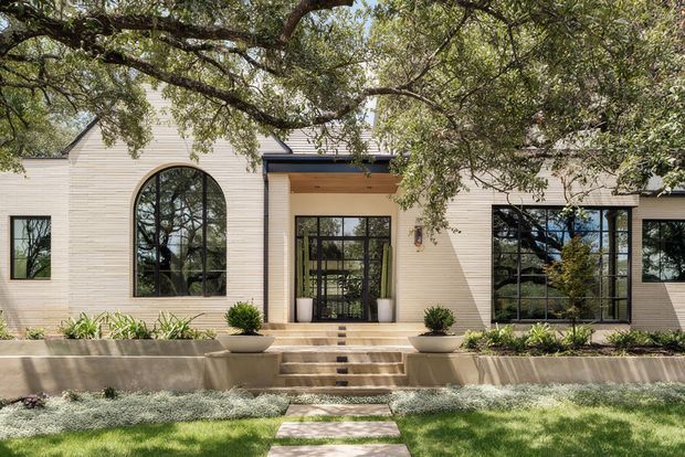 Front exterior view of a low-rise contemporary home with a pale horizontal-clad facade, prominent black-framed glazing, a tall arched window, and a recessed entry beneath a timber-lined canopy. Broad concrete steps and raised planters lead through a landscaped front garden, with mature tree branches framing the facade.