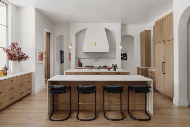 A bright, spacious contemporary kitchen with a large central island and four black bar stools. The room features extensive light timber cabinetry, a white stone-look benchtop, integrated appliances, a sculptural white rangehood, arched wall openings, pendant lights, pale timber flooring, and tall glazing along the left side.