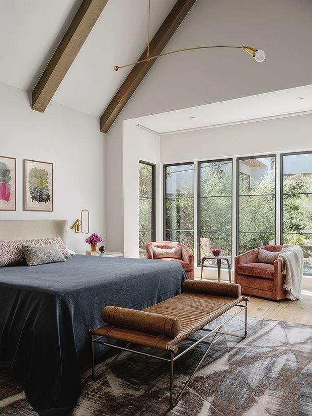 A refined bedroom with a large upholstered bed, dark blue bedding, leather-and-metal bench, two lounge chairs, and a small side table. The room has a high vaulted ceiling with exposed timber beams, pale walls, timber flooring, expansive black-framed glazing, a patterned area rug, framed artwork, and a slim brass ceiling light. The interior is bright, warm, and contemporary with mid-century-inspired furnishings.