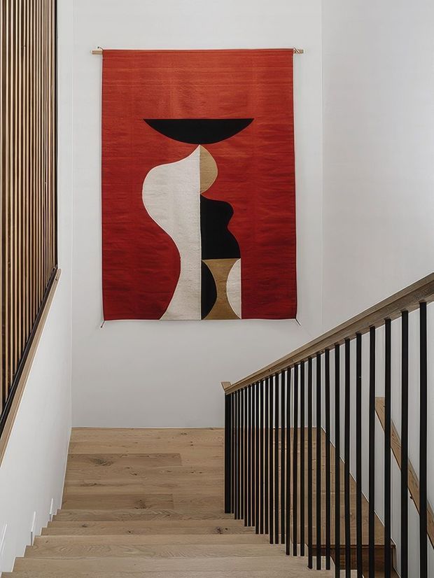Interior view looking up a modern stairway toward a landing, with pale timber stair treads and flooring, slim black vertical balustrade members, and a large red abstract textile artwork mounted on a white wall. A narrow vertical timber slat feature is visible along the left wall, while the stair structure and finishes create a clean, contemporary composition.