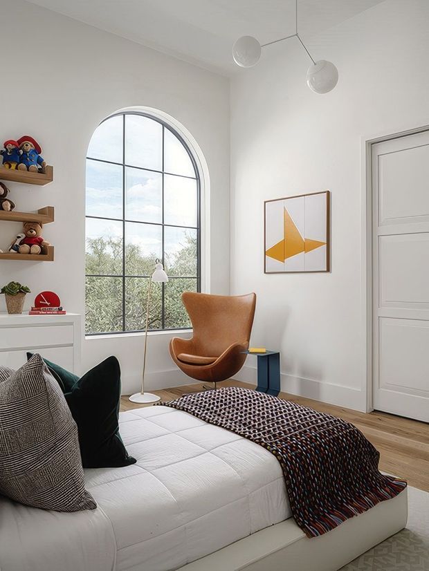 A bright, refined bedroom with a large arched, black-framed window, white walls and ceiling, pale timber flooring, and a low upholstered white bed. A cognac leather lounge chair sits beside a small dark side table, with a sculptural floor lamp, geometric artwork, floating shelves displaying toys, and a textured patterned throw adding character. The room combines clean contemporary architecture with mid-century-inspired furnishings.