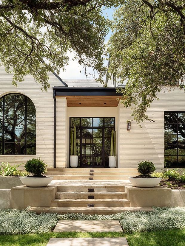 Front exterior of a refined low-rise residence with pale horizontal cladding, a dark-trimmed recessed entry canopy, large black-framed glazed doors and arched windows. Broad stone steps lead through a landscaped garden with clipped shrubs, large white planters and low groundcover, while mature tree branches frame the facade.